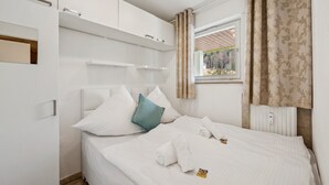 A neatly made bed with white linens, a teal pillow, and a towel, a white wardrobe with a mirror, and a window with curtains.