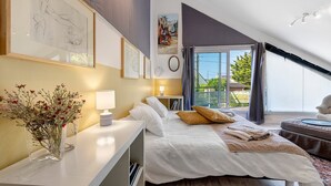 A bedroom with a large bed, a desk with a lamp, a vase with flowers, and a window with a view of greenery.