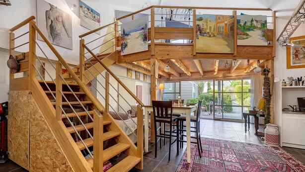 A wooden staircase leading to a loft bedroom with a large window, a dining area, and artwork on the walls.