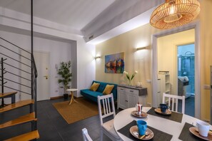 Un interno moderno con una scala, un'area pranzo con un tavolo apparecchiato per due, un divano blu e una cucina visibile attraverso una porta aperta.