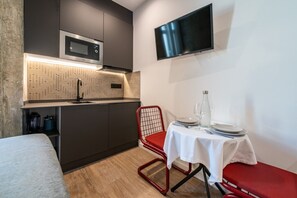 En kompakt tekøkken med mikroovn, vask og flisebeklædt bagvæg. Der er et spiseområde med et bord dækket til to, en rød stol og et tv ophængt på væggen.