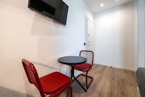 Et lille værelse med et TV ophængt på væggen, et rundt bord med to røde stole og en dør i baggrunden.