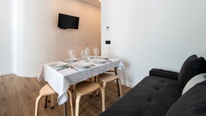 En matplats med ett bord för fyra personer, en platt-TV som är monterad på väggen och en soffa i mörk färg.