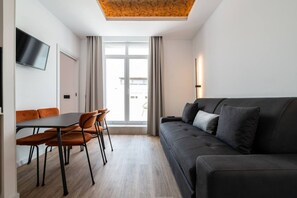 En moderne stue med sofa, spisebord med stole, et fladskærms-TV på væggen og et stort vindue med gardiner.