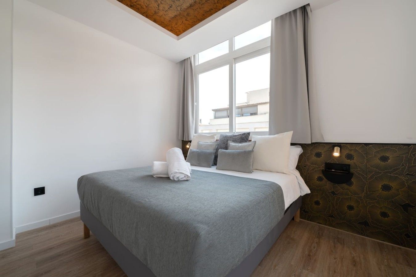 A hotel room with a large bed, a wooden headboard, a window with curtains, and a wall-mounted light fixture.