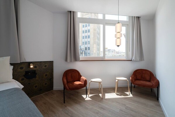 Et moderne hotelværelse med seng, to lænestole, et lille bord, et vindue med gardiner og en hængende lampe.