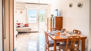 Un coin repas avec une table et des chaises en bois, un téléviseur posé sur un meuble, et un canapé avec des coussins à l’arrière-plan.