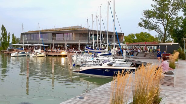 Un port de plaisance avec des bateaux amarrés, un restaurant doté d’une terrasse, et un bâtiment moderne en arrière-plan.