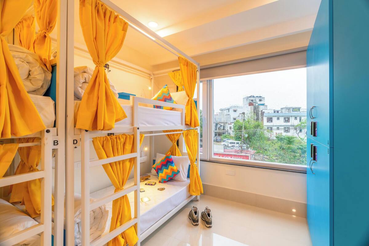 Quarto com beliche, cortinas amarelas, janela com vista para a cidade e um par de sapatos no chão.