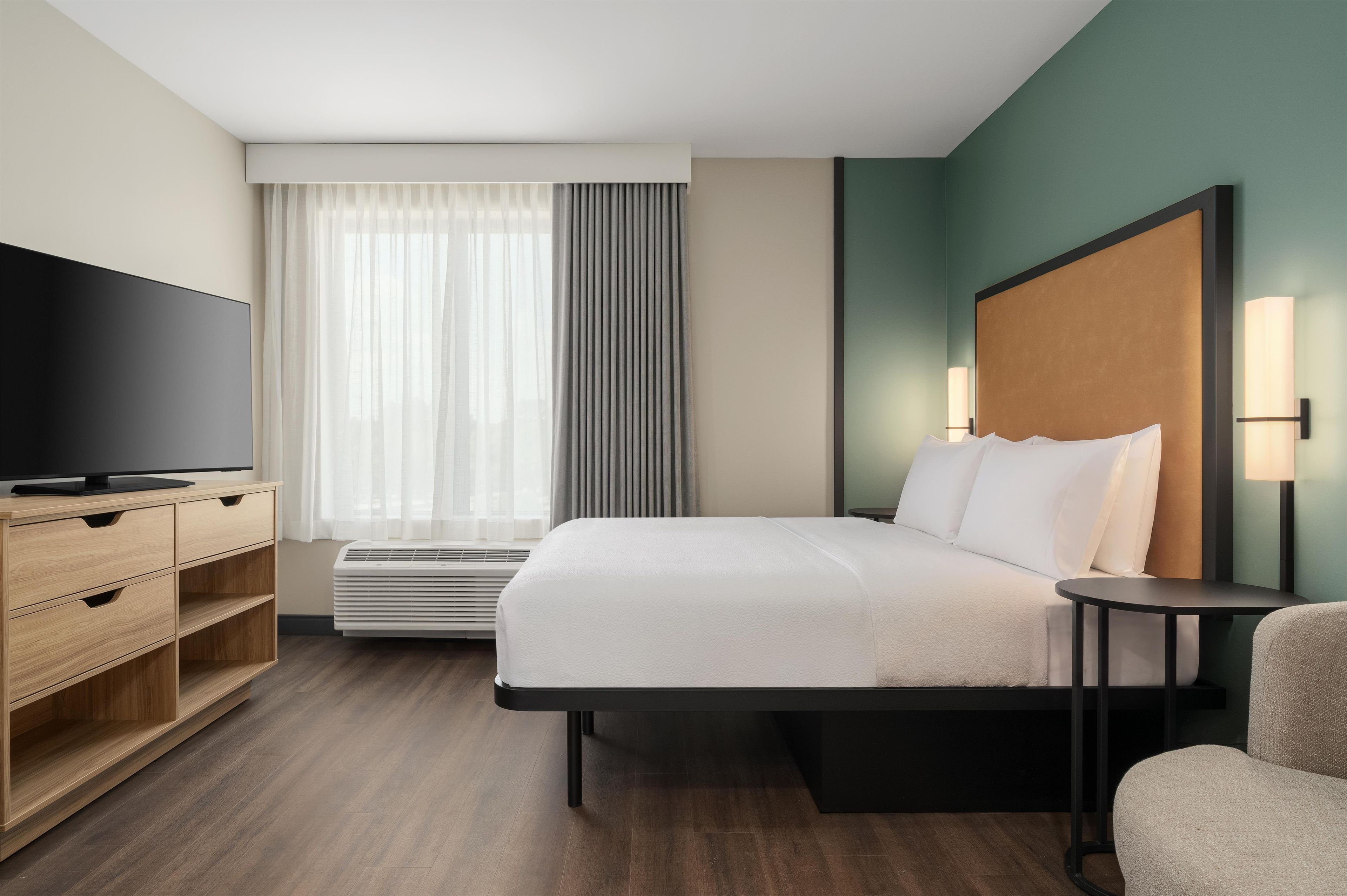 A hotel room with a bed, a TV, a desk, a chair, and a window with curtains.