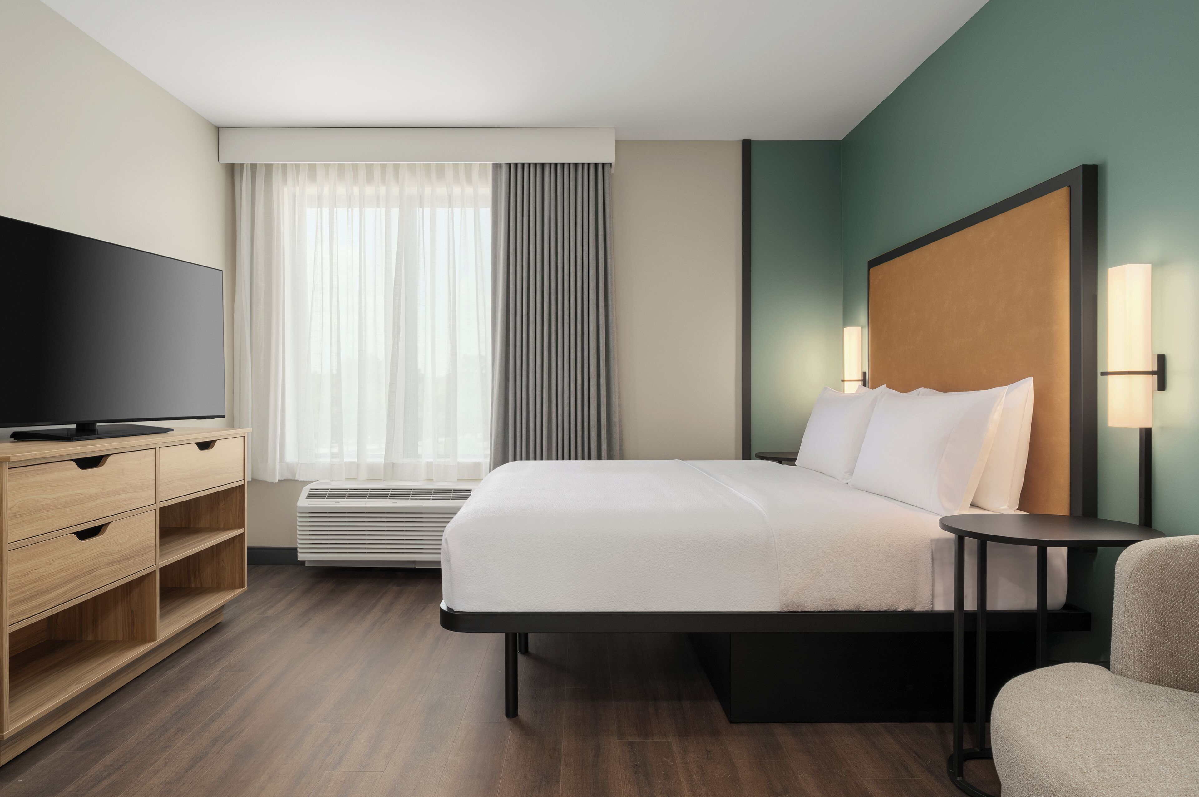 A hotel room with a bed, a TV, a desk, a chair, a window with curtains, and a wall-mounted lamp.