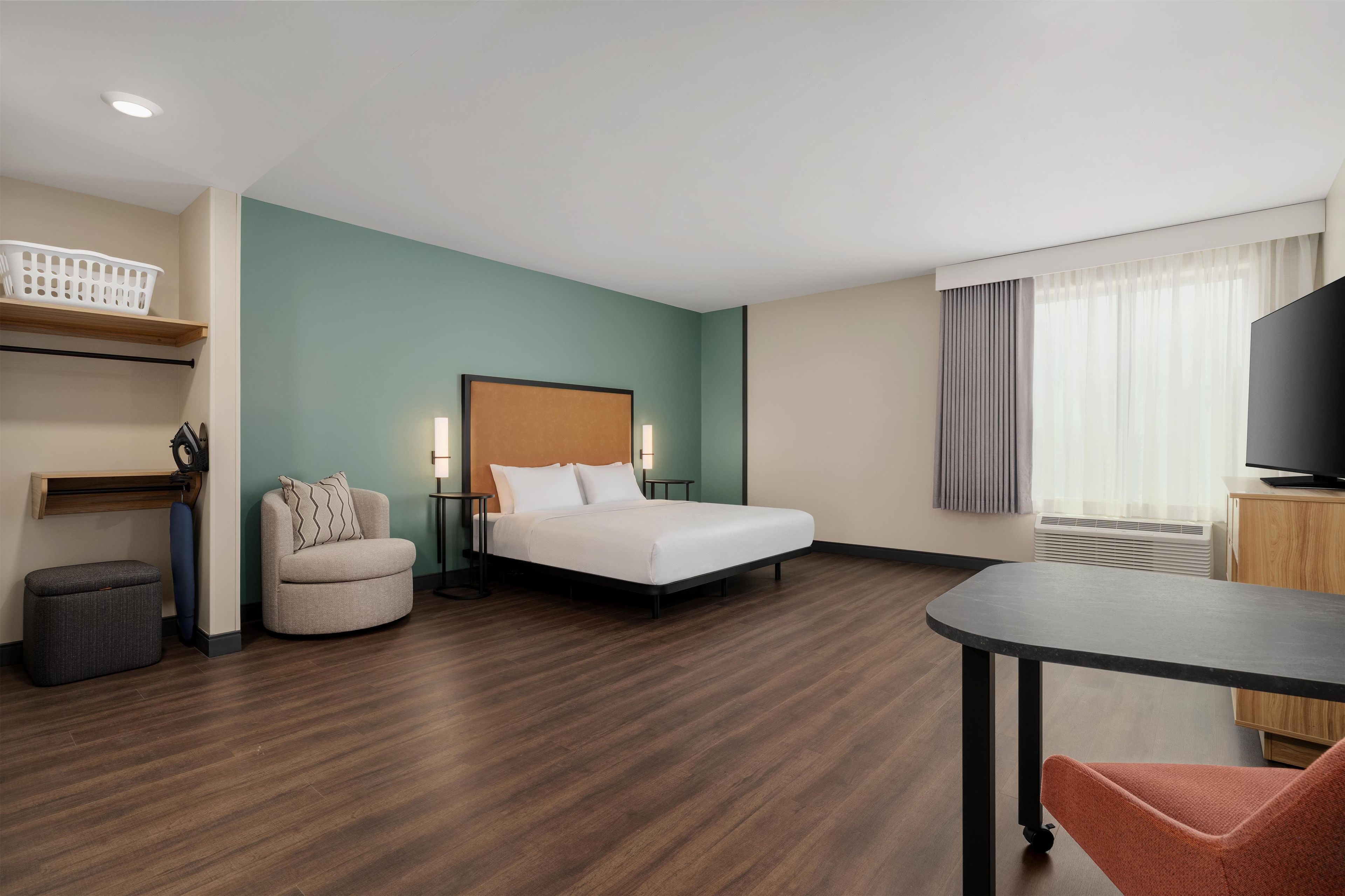 A hotel room with a bed, a chair, a table, a TV, and a window with curtains.