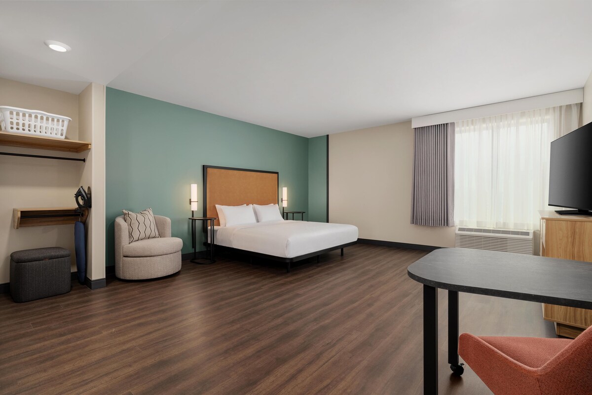 A hotel room with a bed, a chair, a table, a TV, and a window with curtains.