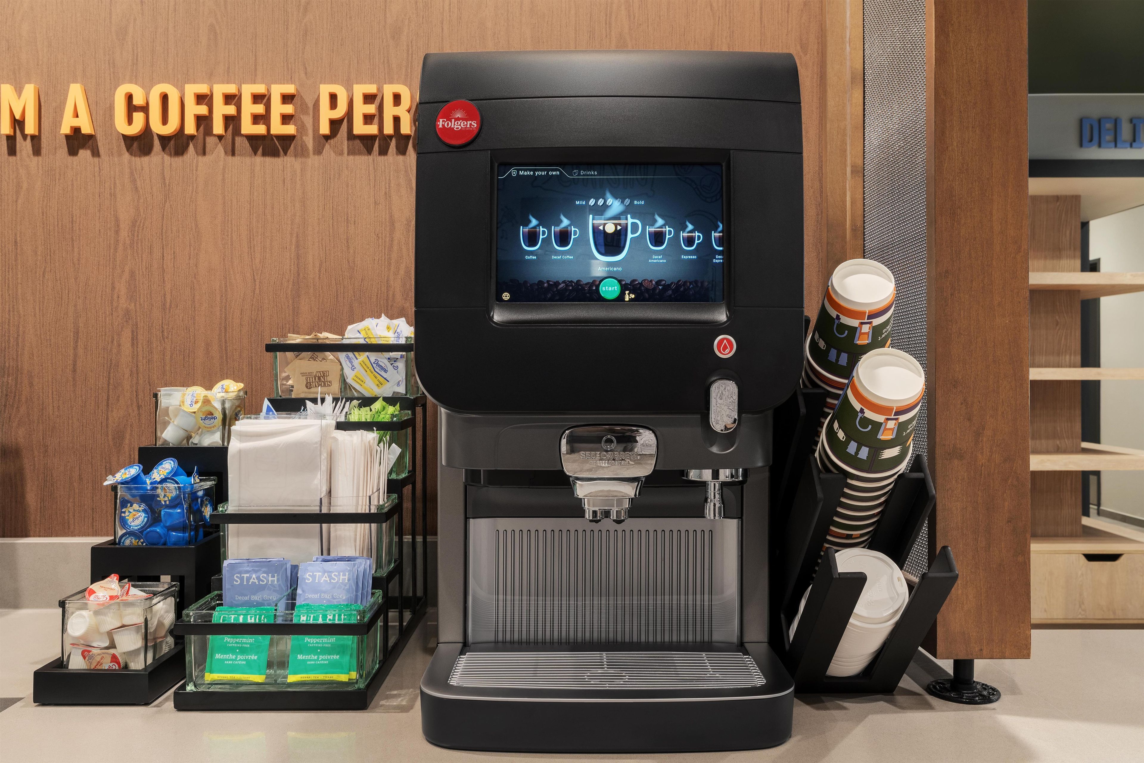 A coffee machine with a digital interface for selecting drinks, a coffee cup holder, and a display of coffee pods and creamer packages.