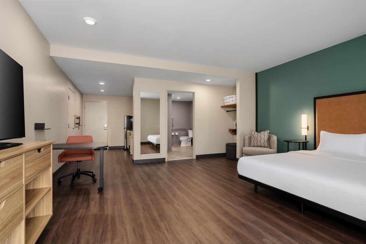 Une chambre d’hôtel avec un lit, un bureau, une chaise et une salle de bain visible à travers une porte ouverte.