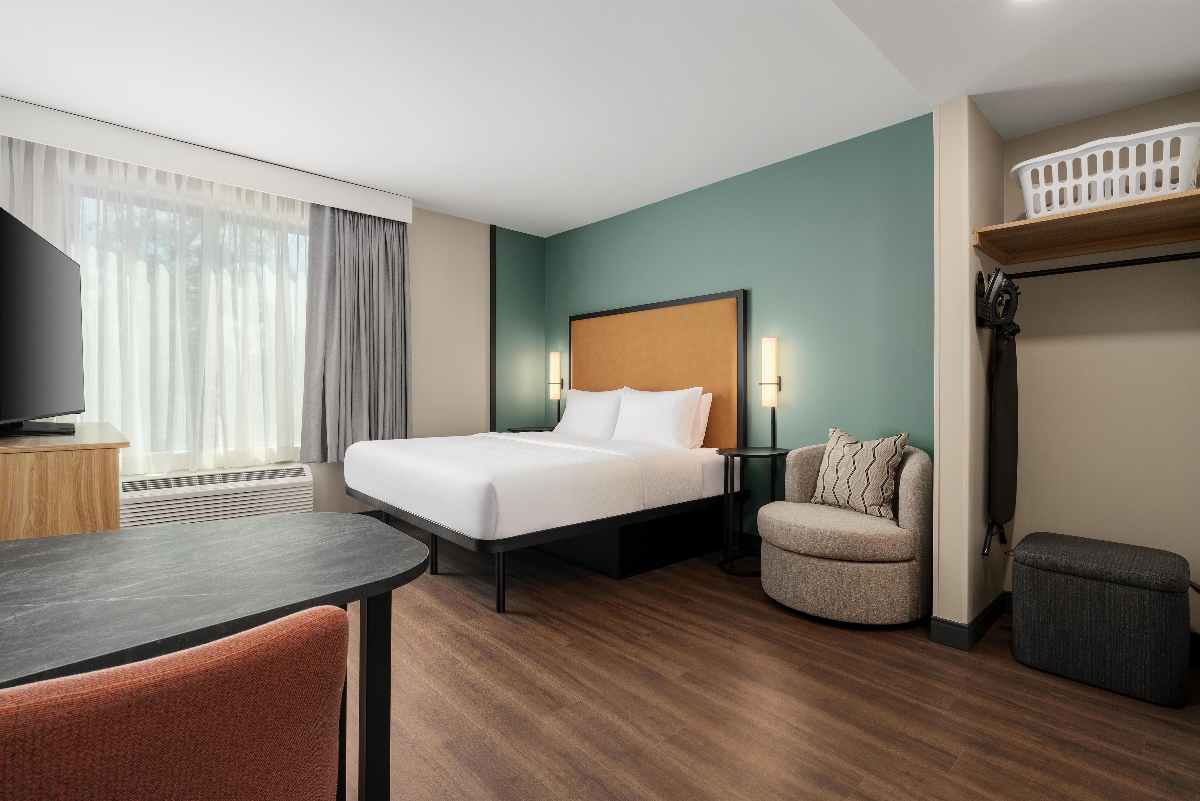 A hotel room with a bed, a desk, a chair, a TV, a heater, a lamp, and a shelf.