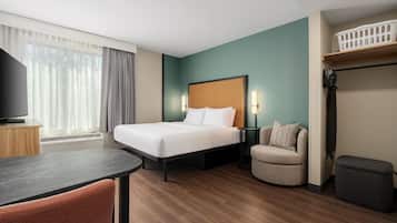 A hotel room with a bed, a desk, a chair, a TV, a heater, a lamp, and a shelf.