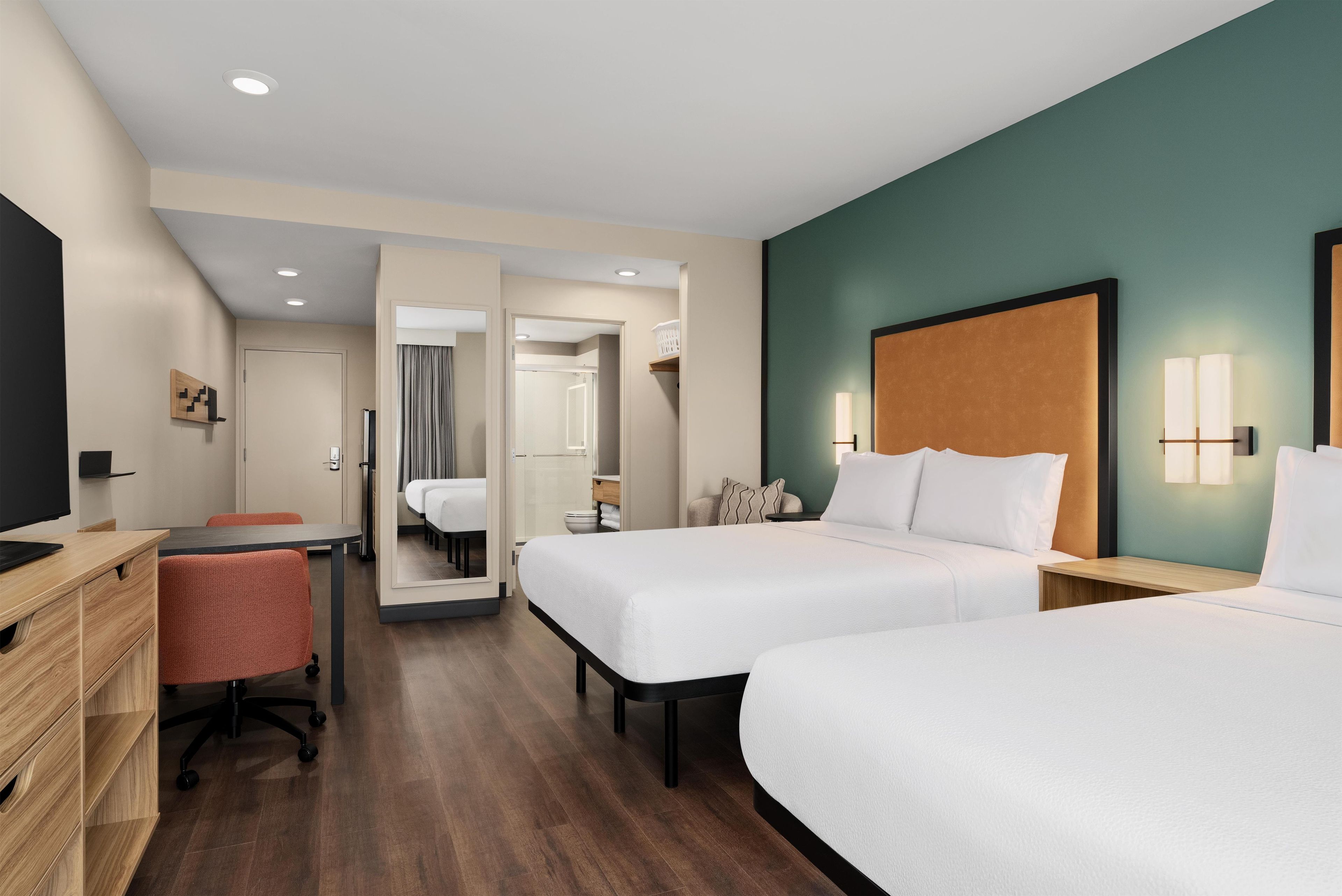 A hotel room with two beds, a desk, a chair, a television, and a bathroom visible through an open door.