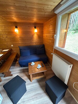 A wooden cabin interior with a sofa, two benches, a small table with cups and a remote control, and a window with a view of trees. - Tipa Lodge Apartment: Mountain View, Pool, Spa & Parking (Cauterets)