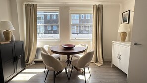 Ein Zimmer mit einem runden Tisch, zwei Stühlen, einem Schrank, einer Lampe und einem fenster mit Vorhängen.