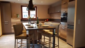 Una cocina moderna con comedor, que incluye una isla central, sillas, una lámpara colgante y un refrigerador.