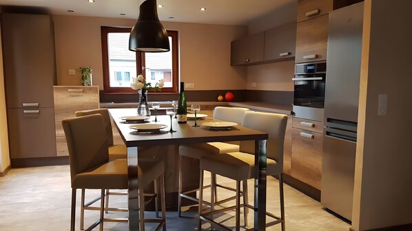 A modern kitchen with a dining area, featuring a central island, chairs, a pendant light, and a refrigerator.