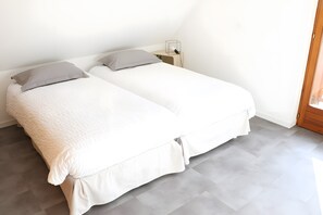 Two single beds with white bedding and grey pillows in a room with a wooden floor and a nightstand with a lamp.