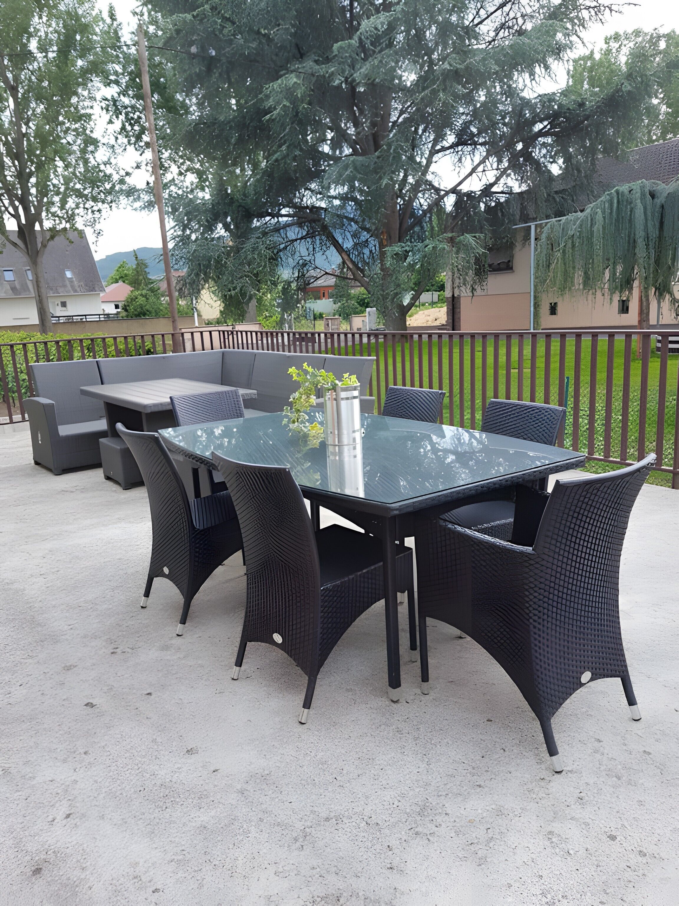 An outdoor patio with a glass-top table surrounded by wicker chairs, a metal vase with greenery, and a wooden fence in the background.
