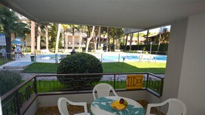Un balcón con mesa y sillas, con vista a una piscina y un área verde con palmeras.