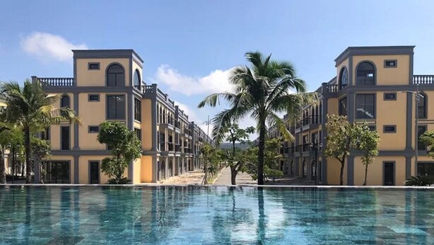 A pool with a clear blue surface, surrounded by buildings with a uniform architectural style, palm trees, and a clear sky. - Canisa Villas (Phu Quoc)