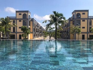 A pool with a clear blue surface, surrounded by buildings with a uniform architectural style, palm trees, and a clear sky. - Canisa Villas (Phu Quoc)