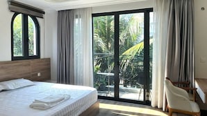 A hotel room with a large bed, a desk, a chair, and a sliding glass door leading to a balcony with plants. - Canisa Villas (Phu Quoc)