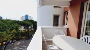 A balcony with a table and chairs, a window, and a view of trees and buildings. - Beachside and equipped apartment (Lignano Sabbiadoro)