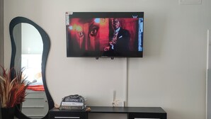A modern living room with a flat-screen TV mounted on the wall, a round glass coffee table, a black console table with decorative items, and a large mirror. - Modern Cosy Studio in Ruaka Near Two Rivers Fast WiFi and fParking,24/7 Security (Nairobi)
