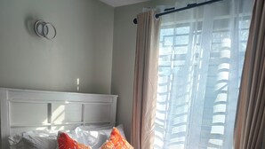 A neatly made bed with pillows and a blanket, a bedside table, a window with curtains, and a wall clock. - Modern Cosy Studio in Ruaka Near Two Rivers Fast WiFi and fParking,24/7 Security (Nairobi)