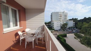 Varanda com cadeiras e mesa de plástico branco, com vista para um estacionamento e um edifício de vários andares.