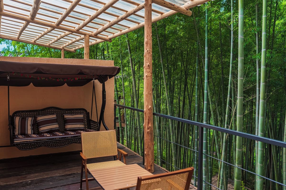 Un balcón con un banco de madera, una silla de mimbre y una mesa. Un bosque de bambú al fondo.