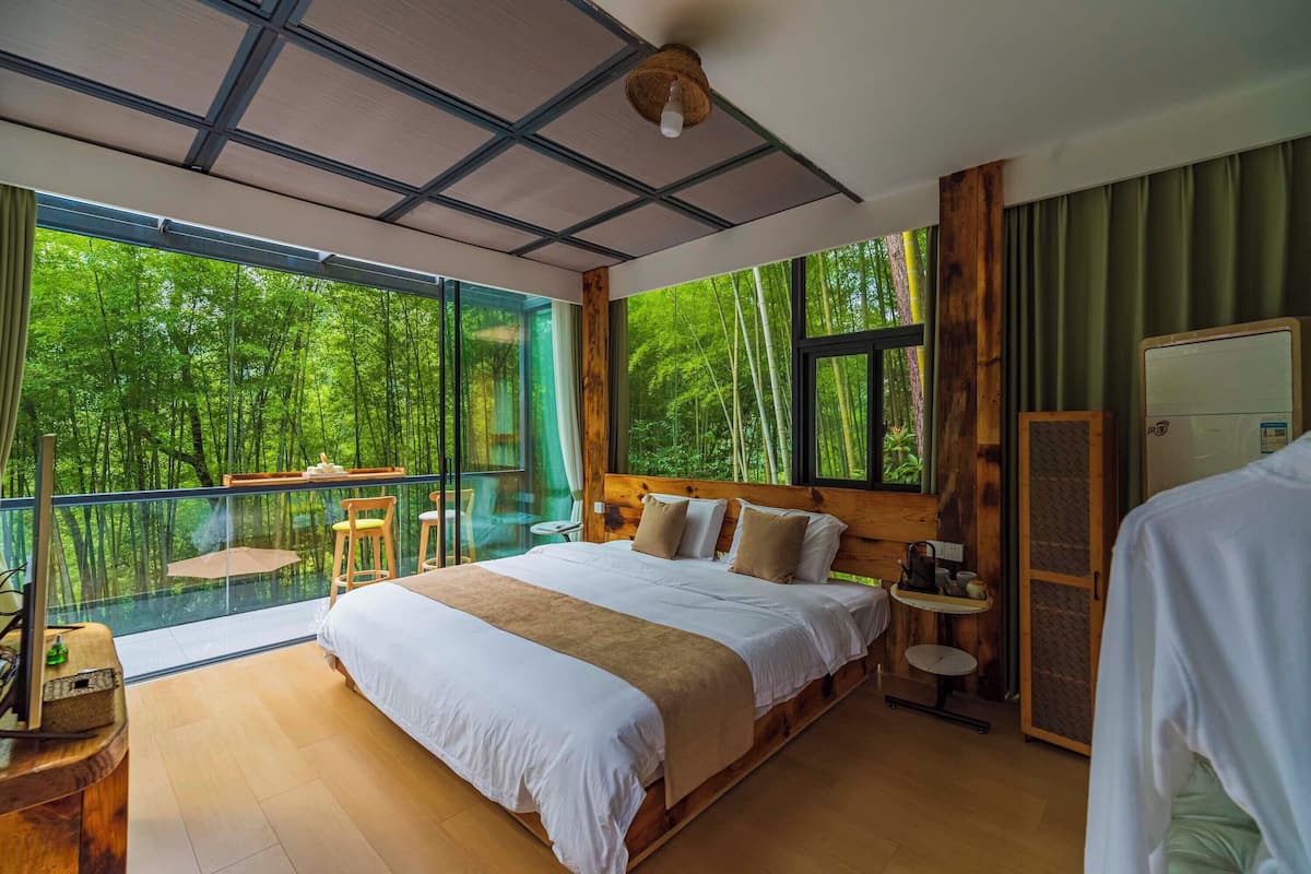 Un dormitorio con una cama grande, una mesita pequeña, una silla y vistas a un bosque a través de amplios ventanales.