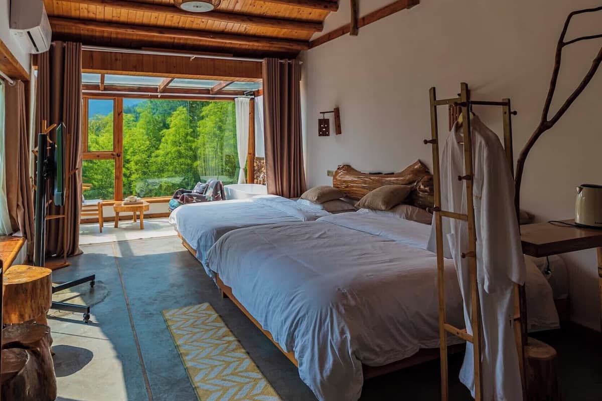 Un dormitorio con techo de madera, una cama, una mesita de noche, un perchero y una ventana con vistas a árboles.