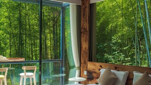 Ein Schlafzimmer mit einem großen Bett, einem hölzernen Kopfteil, einem Glastisch mit Stühlen und Blick auf einen Wald durch große Fenster.