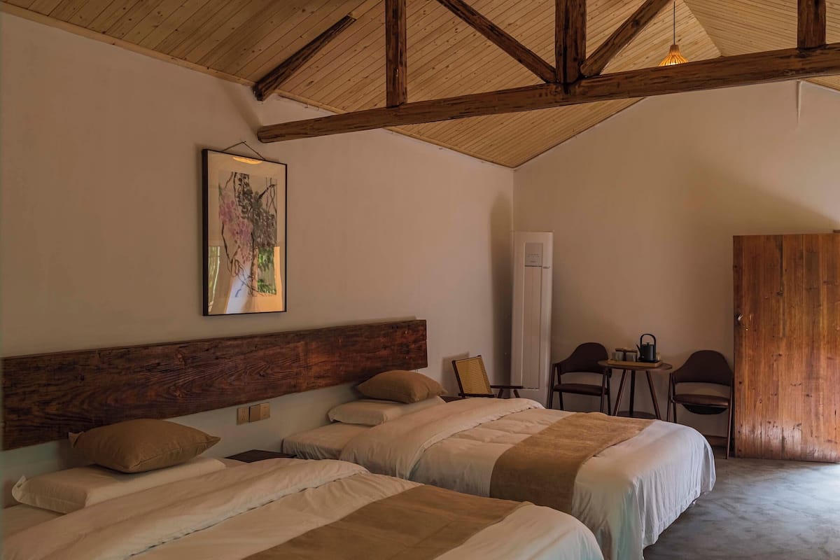 Un dormitorio con dos camas, techo de madera, un cuadro, una mesita con sillas y un armario.