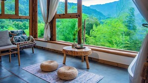 Ein gemütliches Zimmer mit einem Holzstuhl, einem kleinen runden Tisch mit einer Pflanze und Blick auf üppiges Grün durch große Fenster.