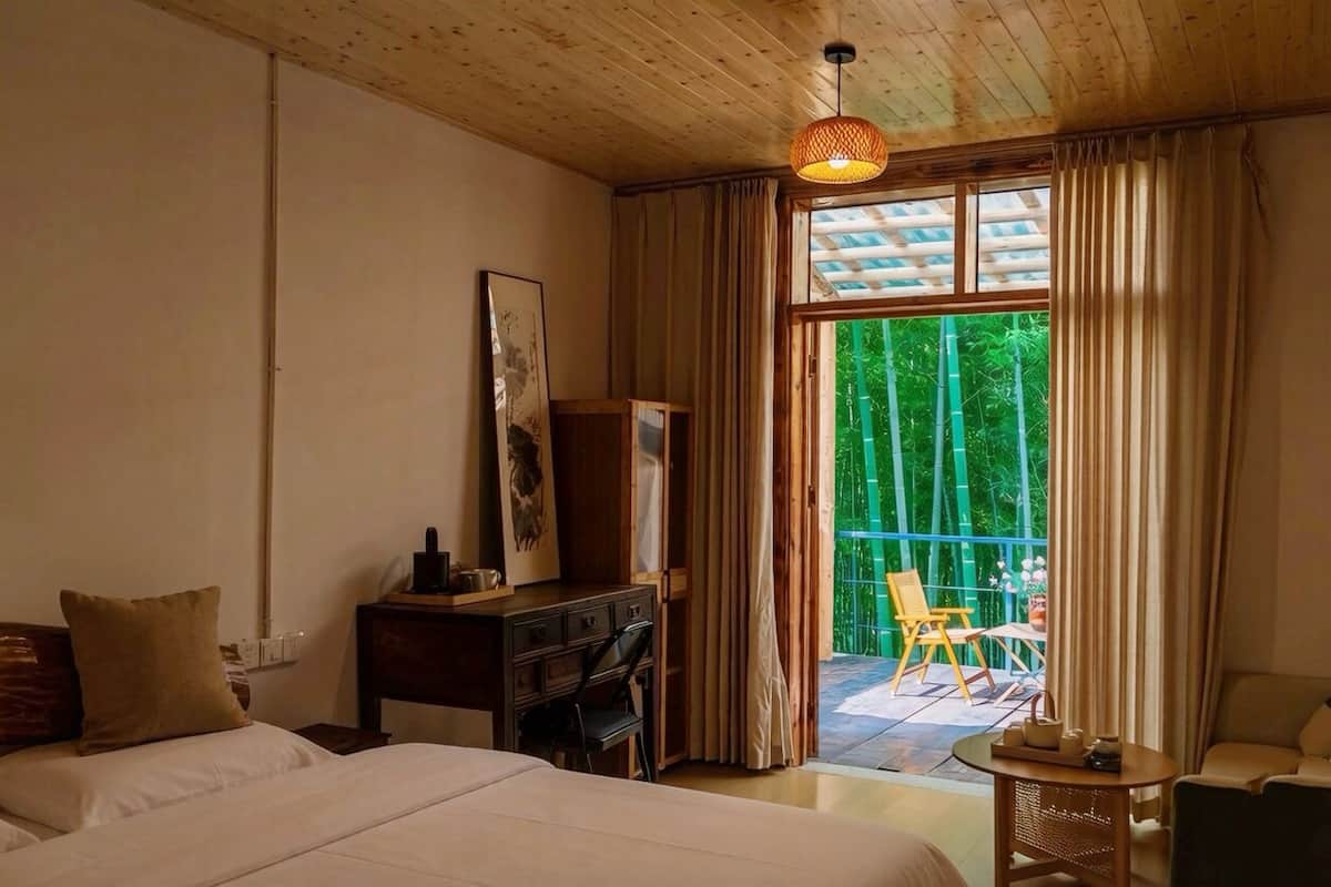 Un dormitorio con cama, escritorio, silla y balcón con vista a zonas verdes.