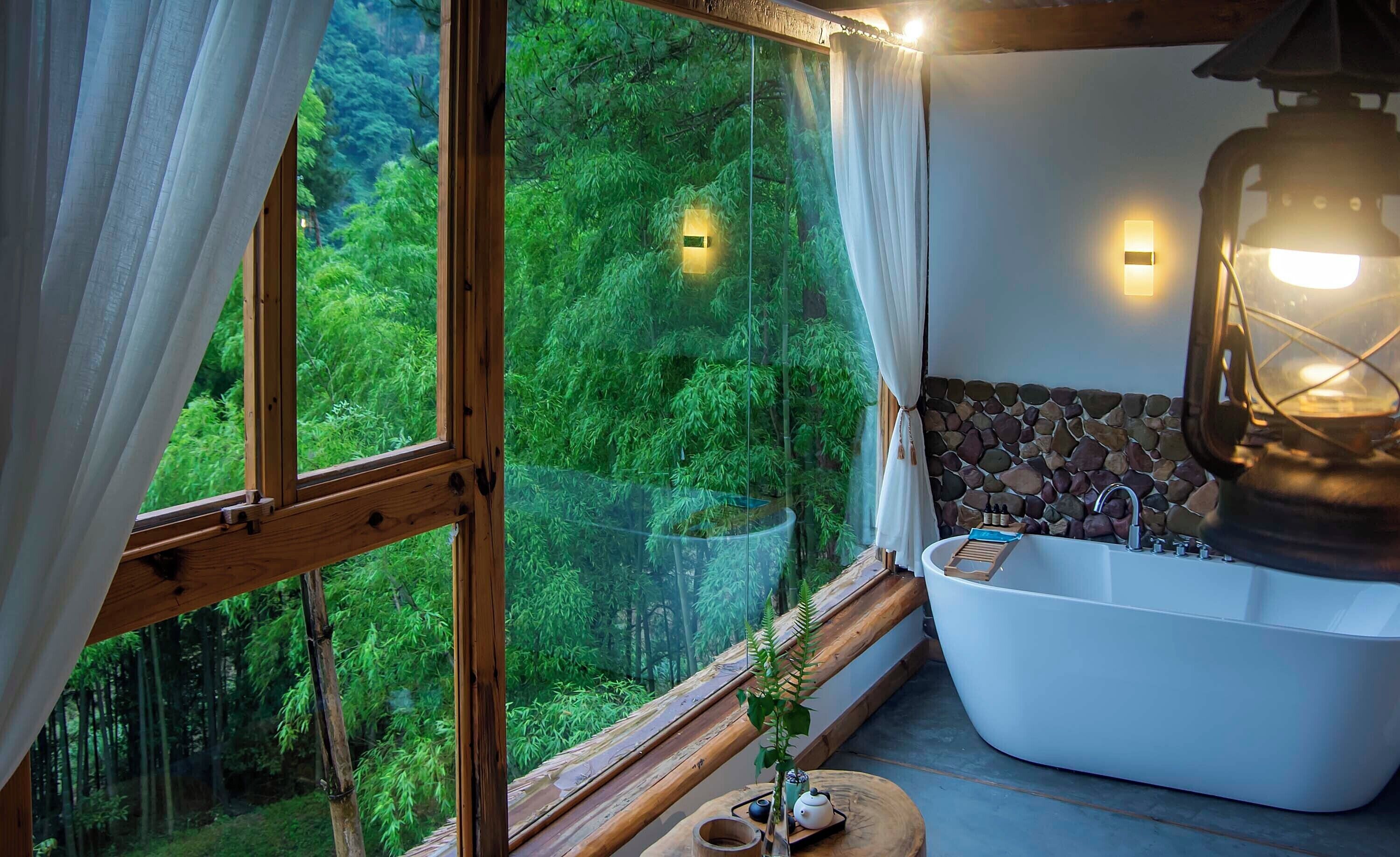 A bathroom with a bathtub, a stone wall, and a view of a forest through large windows.