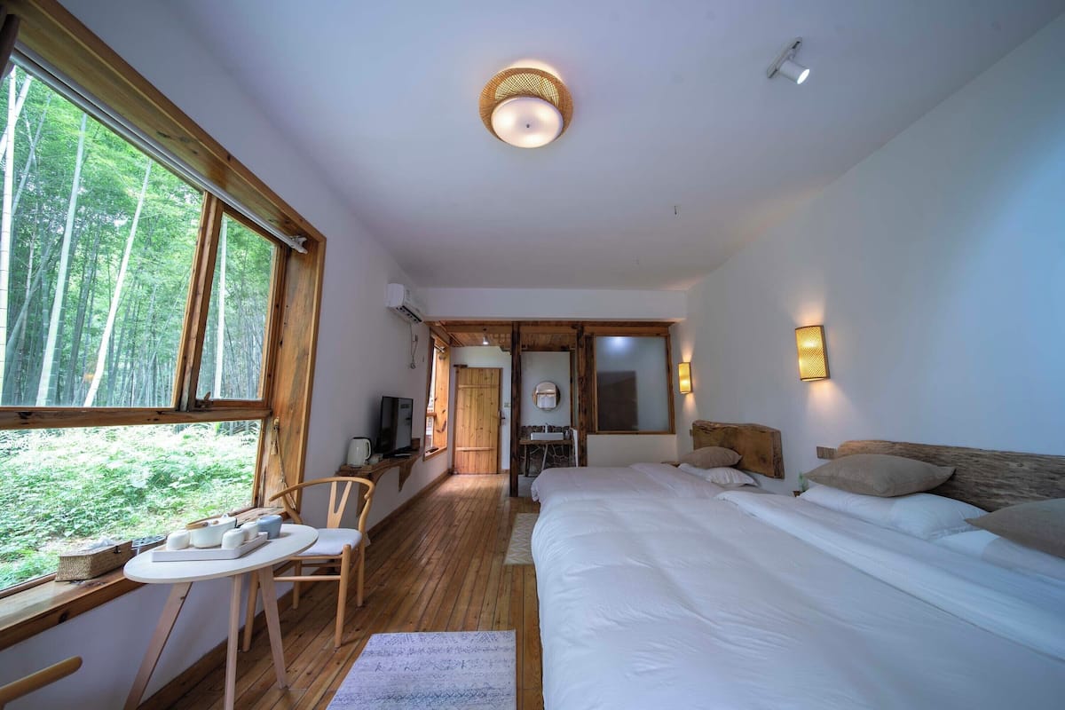 Un dormitorio con una cama grande, un escritorio de madera, una silla, una mesita y una ventana con vistas a árboles.
