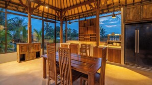 An open-plan kitchen with wooden cabinets, a dining area with chairs, and large windows offering a view of palm trees. - Suasana Harmonis Private Villa (Ubud)