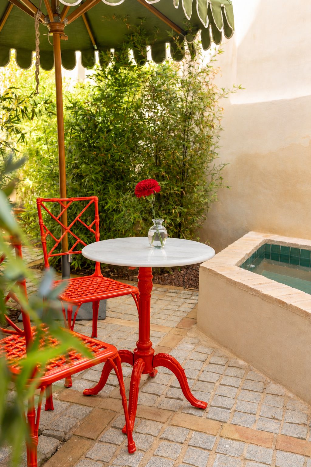 Un patio con una mesa redonda y sillas rojas, un jarrón pequeño con una flor y una piscina al fondo.