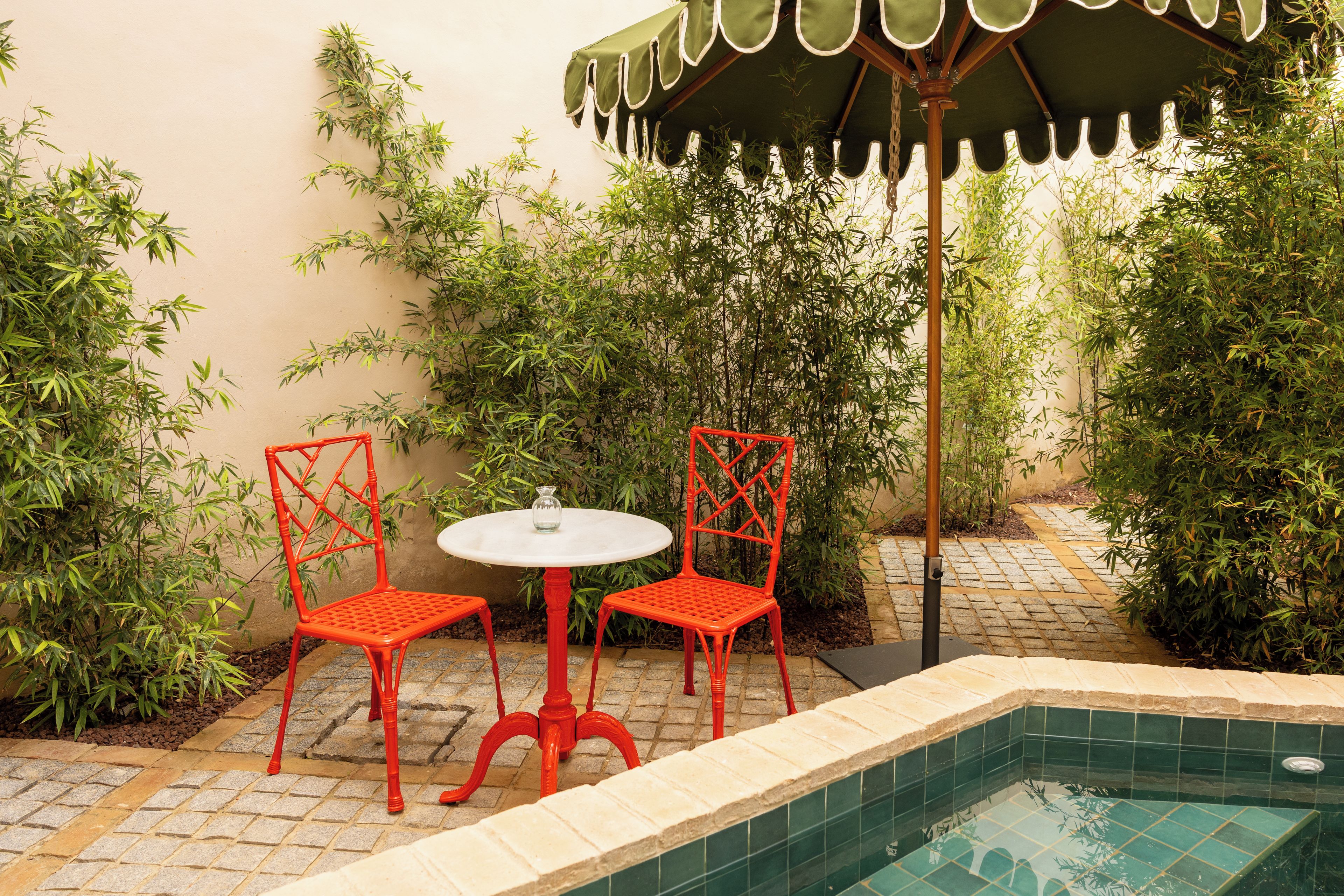 Un pequeño espacio al aire libre con dos sillas rojas y una mesa blanca, rodeado de abundante vegetación y una piscina con azulejos.