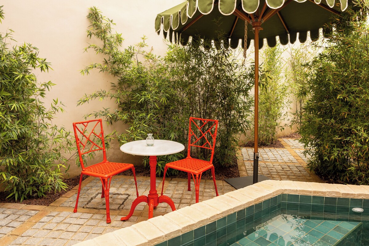 Un pequeño espacio al aire libre con dos sillas rojas y una mesa blanca, rodeado de abundante vegetación y una piscina con azulejos.