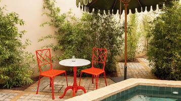 Un pequeño espacio al aire libre con dos sillas rojas y una mesa blanca, rodeado de abundante vegetación y una piscina con azulejos.